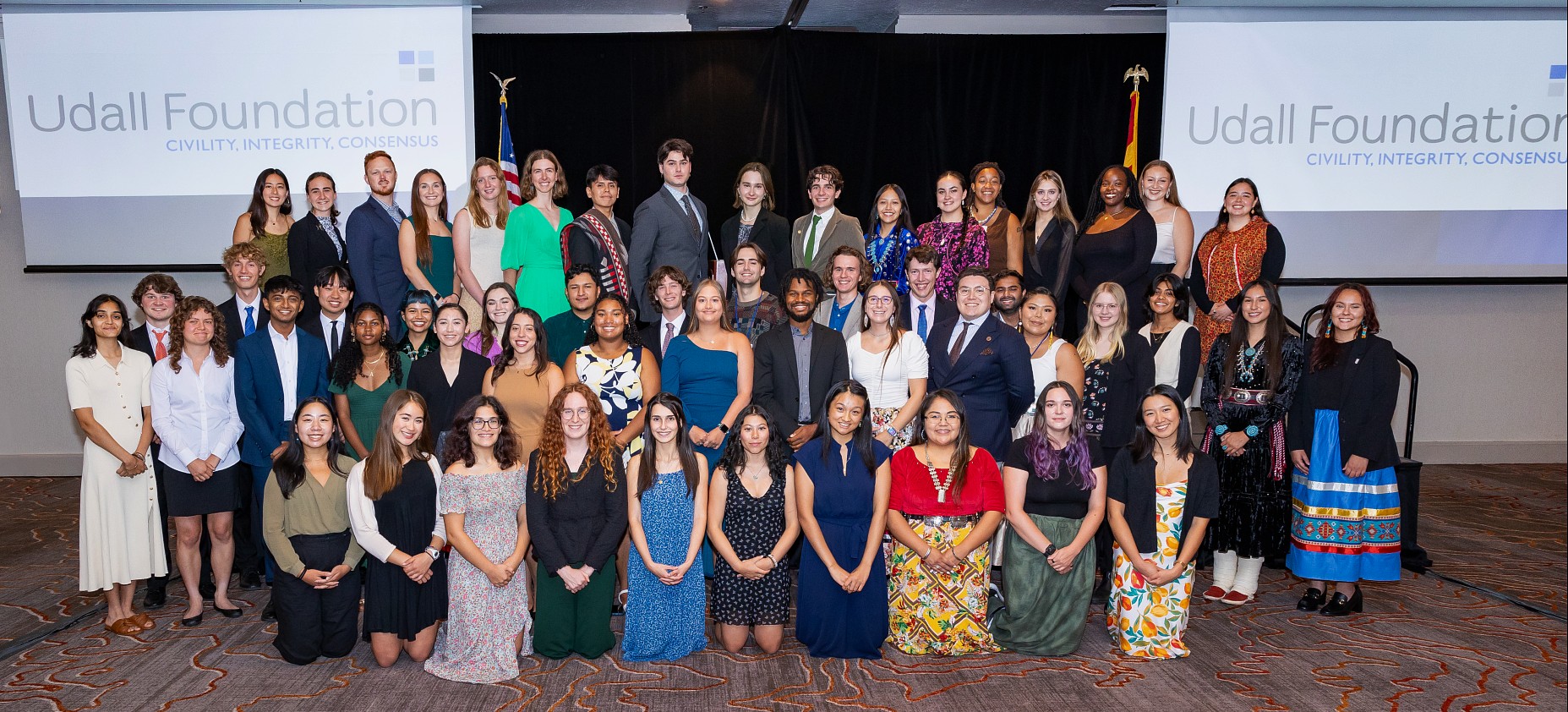 A diverse group of people pose in formal attire at the Udall Foundation event. Two screens display the foundation’s name. The mood is celebratory.