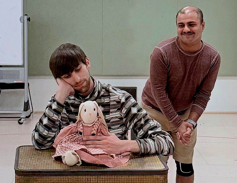 In a brightly lit rehearsal room with pale green walls, a man sits behind a stack of vintage suitcases, leaning his head on his hand while holding a plush toy rabbit wearing a pink dress, while another actor stands to the right, watching the scene with a smiling and amused expression.