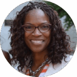 A smiling woman with curly hair and glasses is wearing a white blazer and striped shirt. The background is blurred greenery.