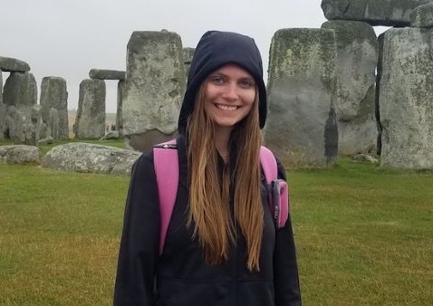 Woman in a black jacket with the hood on smiling at the camera in front of Stonehenge