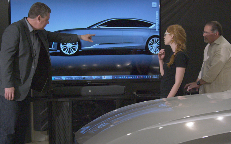 A man in a suit points to a screen showing a silver model of a car. A woman and another man stand to his right alongside the actual car