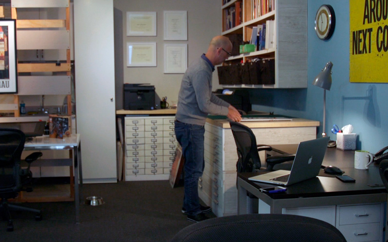 Bald man in gray sweater standing above file drawers and a work surface in a design studio
