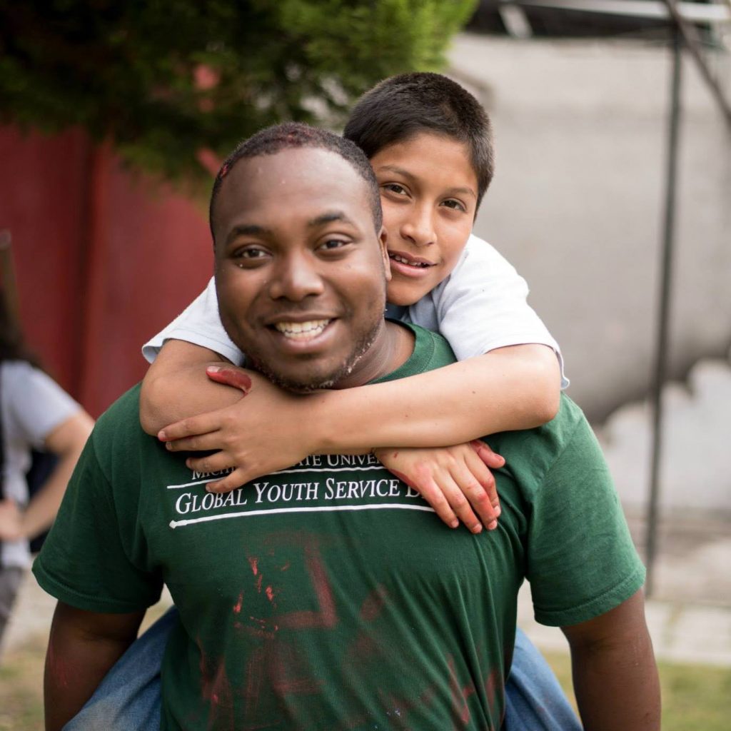man wearing a green shirt with a child wrapping his arms around the man's and hanging on the back