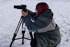 woman holding a camera with a jacket on, she is crouched down on the snowy ground