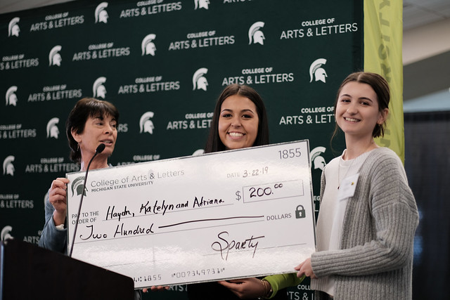 Three women hold a big check. Woman on left has short black hair, woman in the middle has brown hair, and woman on the right has brown hair and is wearing a gray sweater.