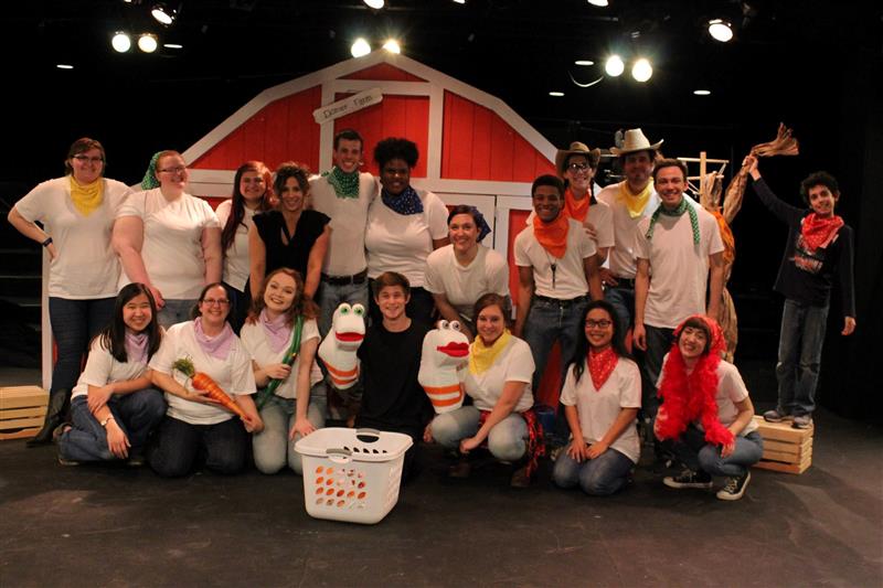 Cast of a theatre show. They all wear white t-shirts and are standing in front of a barn door