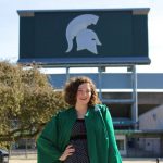 Woman wearing green graduation gown over a black dress standing in front of the spartan stadium