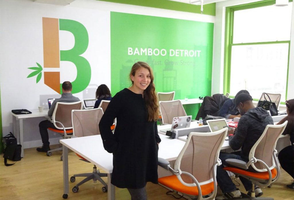 Woman with long brown hair wearing a black long sleeve dress standing in an office with a desk and chairs and a white, orange, and green wall that says "Bamboo Detroit" on it