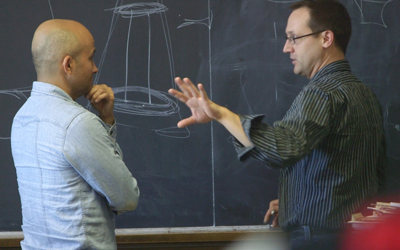 Two men standing in front of a chalkboard. One is explaining to the other what the circular drawing means.