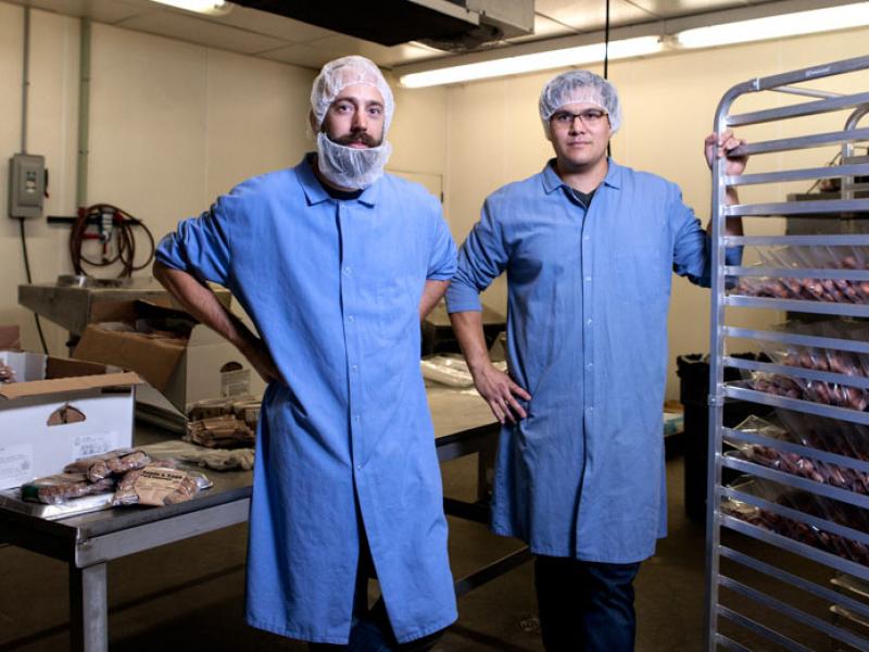 two men who are wearing long blue aprons and hair nets who are standing in a room with packaged meat around them