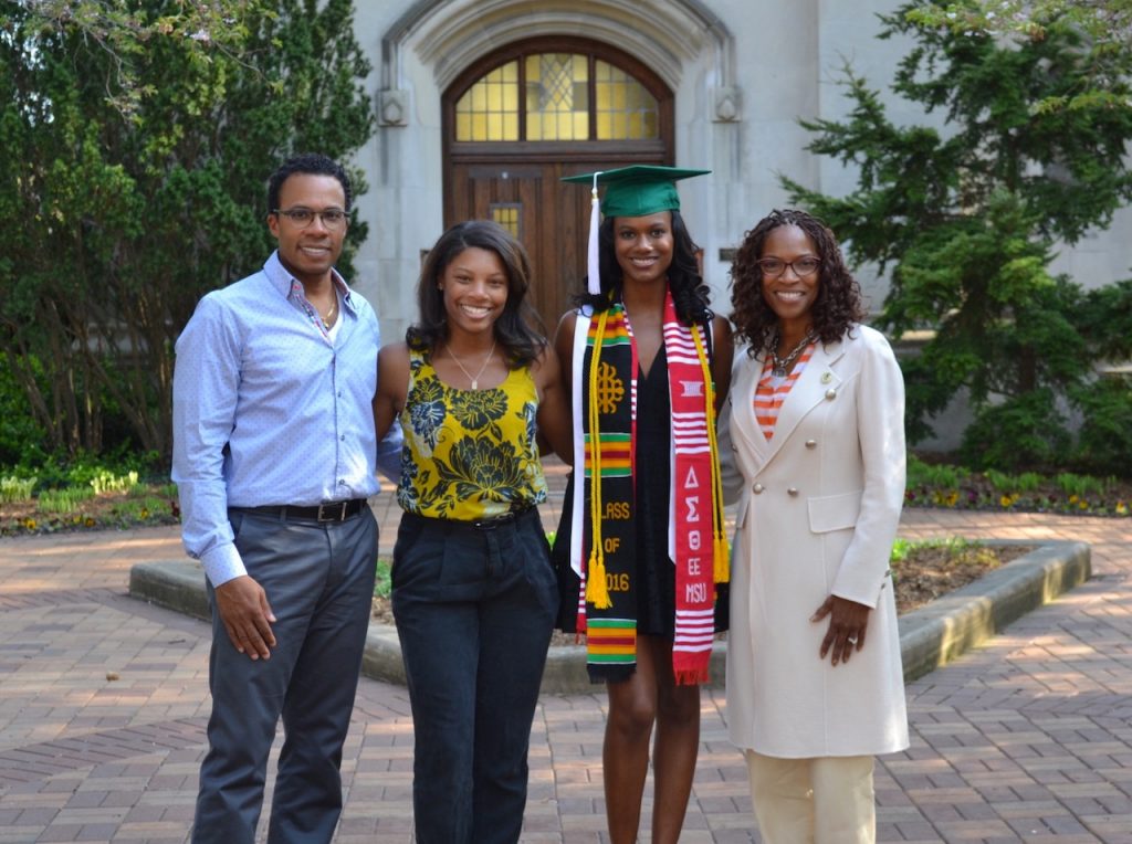 Family of four posing for photo posing in front of a historic building and one of the daughters is wearing a graduation cap 