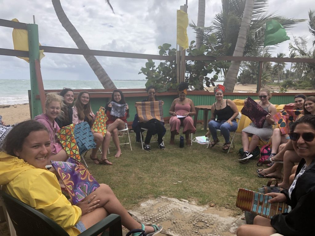 Group of women sitting in a circle by the beach. They each have a square piece of fabric with varying designs, which will be made into head wraps.