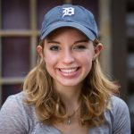 Photo of girl with short blonde hair smiling wearing a grey short sleeve shirt, a silver necklace, and a Detroit Tigers navy baseball cap