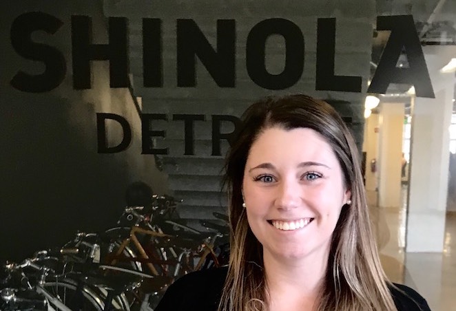 Photo of person wearing a black shirt with a sliver necklace smiling in front of a glass wall that says Shinola Detroit