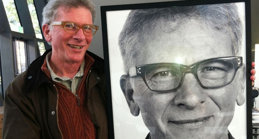 Photo of man smiling with glasses wearing a red sweater and a brown jacket holding a black and white photo of himself 
