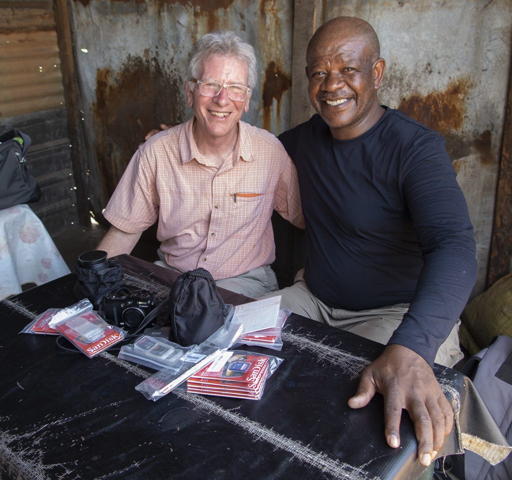 Photo of two men smiling sitting next to each other with their arms around one another with the left wearing an orange checkered shirt and grey pants and glasses and the right wearing a navy shirt and grey pants