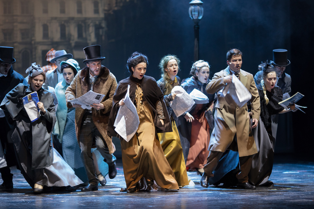 a group of six women and four men on a stage, who are all wearing neutral, old fashioned clothing who are running forward and look as if they are singing 