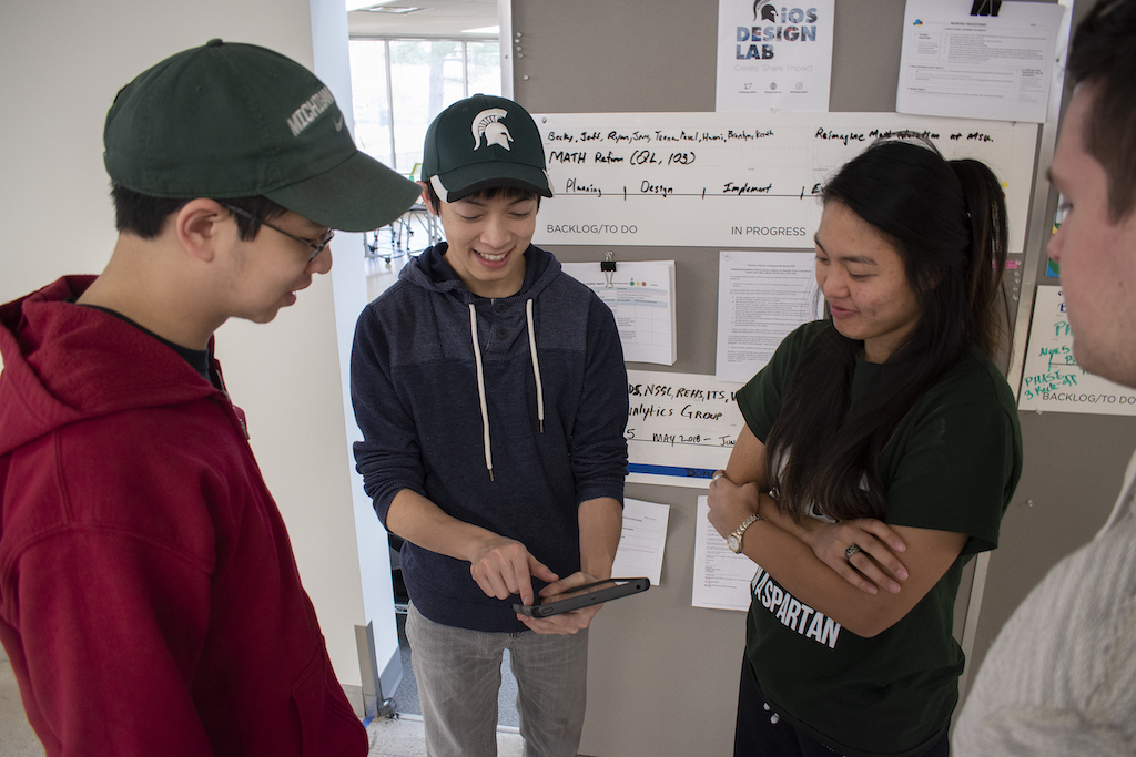 photo of four students, who all have dark hair and are looking at a small tablet that one of them is holding