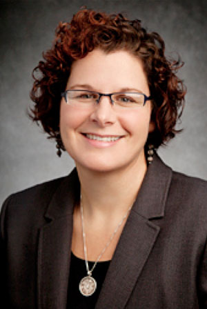 a women with curly hair wearing glasses and a gray blazer with a black shirt and a necklace 