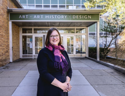 a women with short hair and glasses with a pink and purple scarf and black dress standing in front of an art building 