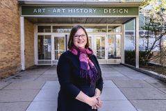 a women with short hair and glasses with a pink and purple scarf and black dress standing in front of an art building