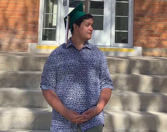 man in a blue button-up standing in front of a brick building with a graduation cap