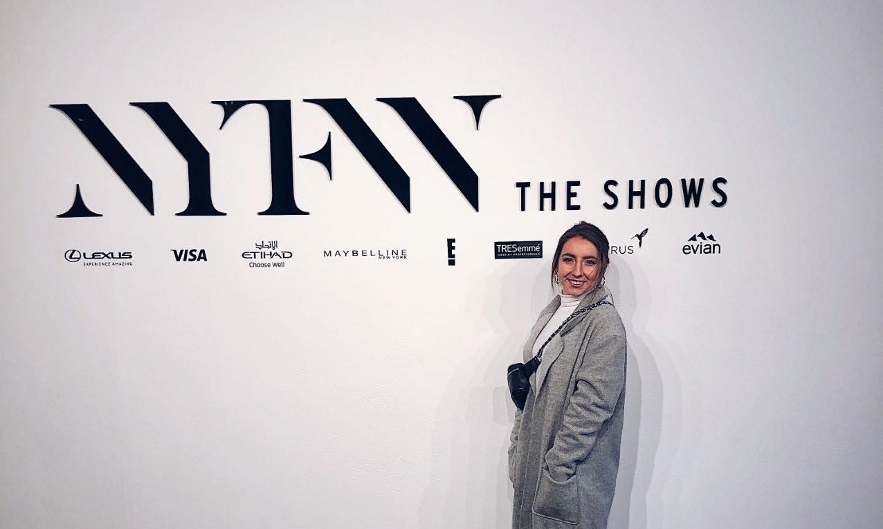 Photo of person wearing grey coat with white shirt and black purse standing in front of white wall that says 'NYFW The Shows'