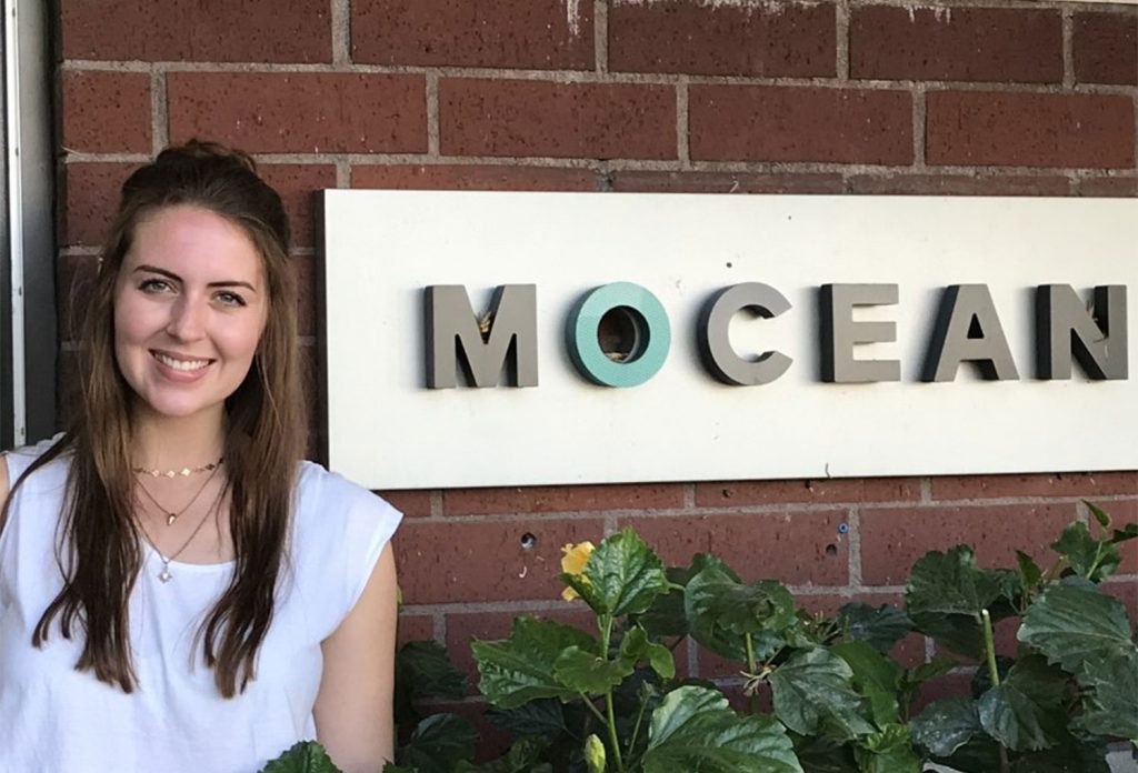 Girl with long brown hair smiling standing next to a sign that says "Mocean"