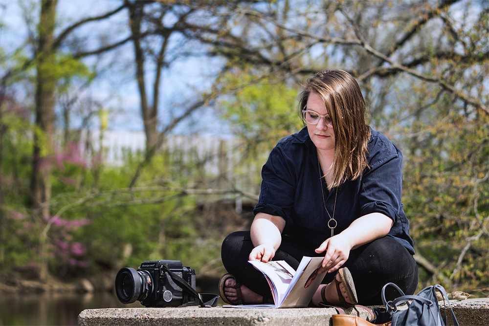 Woman with long brown hair and glasses sitting next to a river with a book and a camera