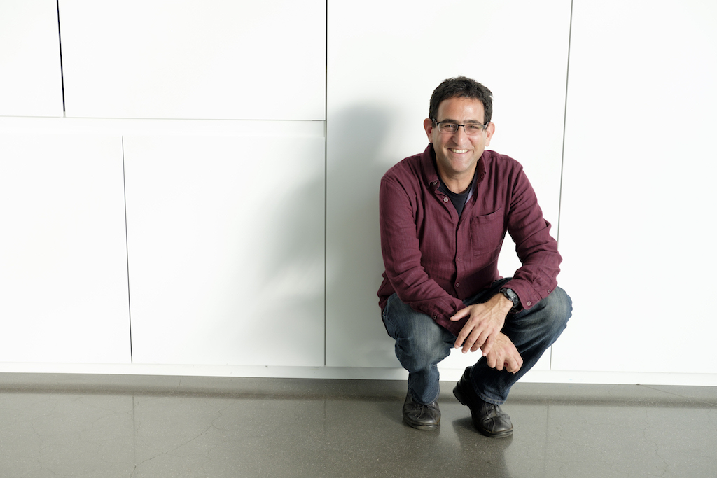 man with short brown hair who is wearing black glasses and wearing a button-down dark red shirt, jeans, and black shoes and is crouching on the ground in front of a white wall