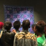 back of a group of six people's heads looking at a purple and blue quilt that is hanging on the wall