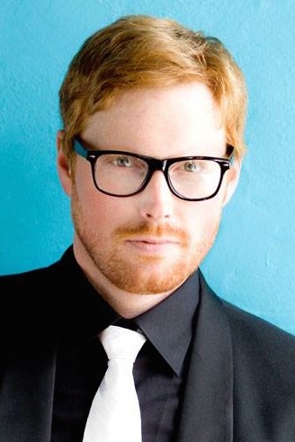 headshot of a man in a black suit and white tie. He has orange hair and beard as well as black glasses