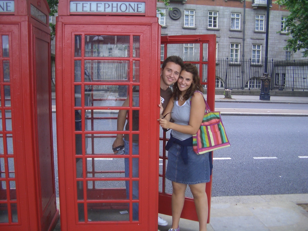 A man and a woman smiling, standing in a red telephone booth