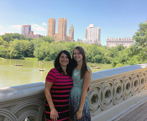 woman with straight, long, brown hair standing next to a shorter woman with medium-length black hair on a bridge. in the background there are tall buildings