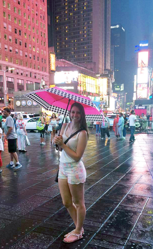 a woman with long, brown hair standing in NYC time square while holding an umbrella