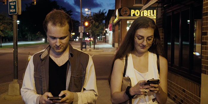 man and woman walking side by side, both are looking down at their phones