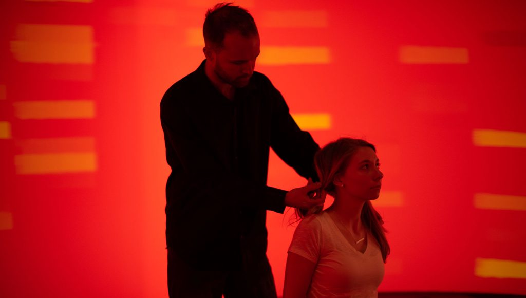 Man standing in front of a red background tying a woman's hair up