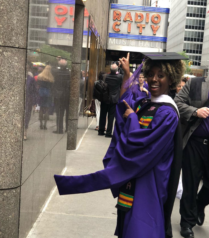 Woman wearing a purple graduation gown and black graduation cap pointing at a Radio City Music Hall sign