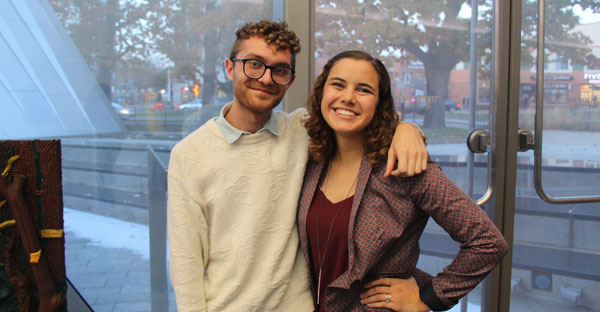 photo of a man and woman smiling and posing for the camera. the man(left has curcly hair and is wearing a button-up and sweater and has his arm around the woman who has curly hair and is wearing a top and blazer
