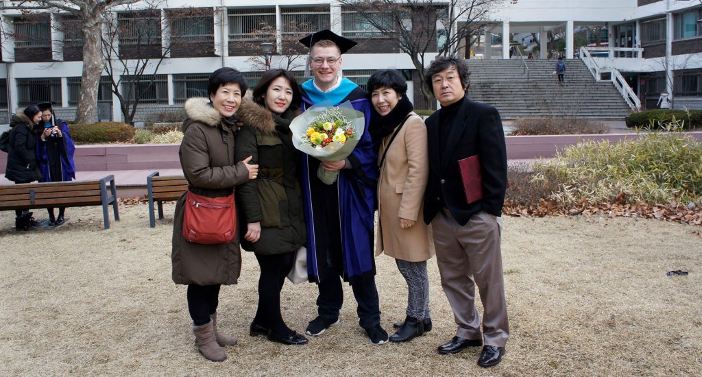Group of 5 people, the man in the middle is wearing a blue graduation cap and gown