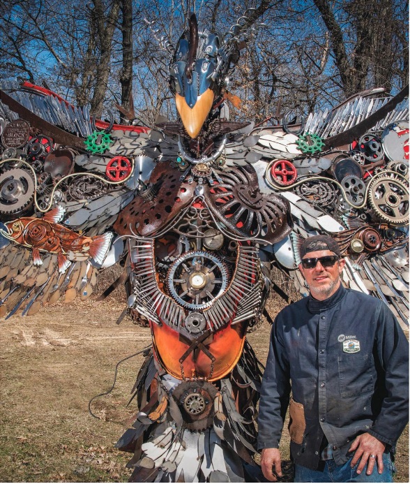 man in sunglasses and denim clothing posing with his falcon-like sculpture made of metal work