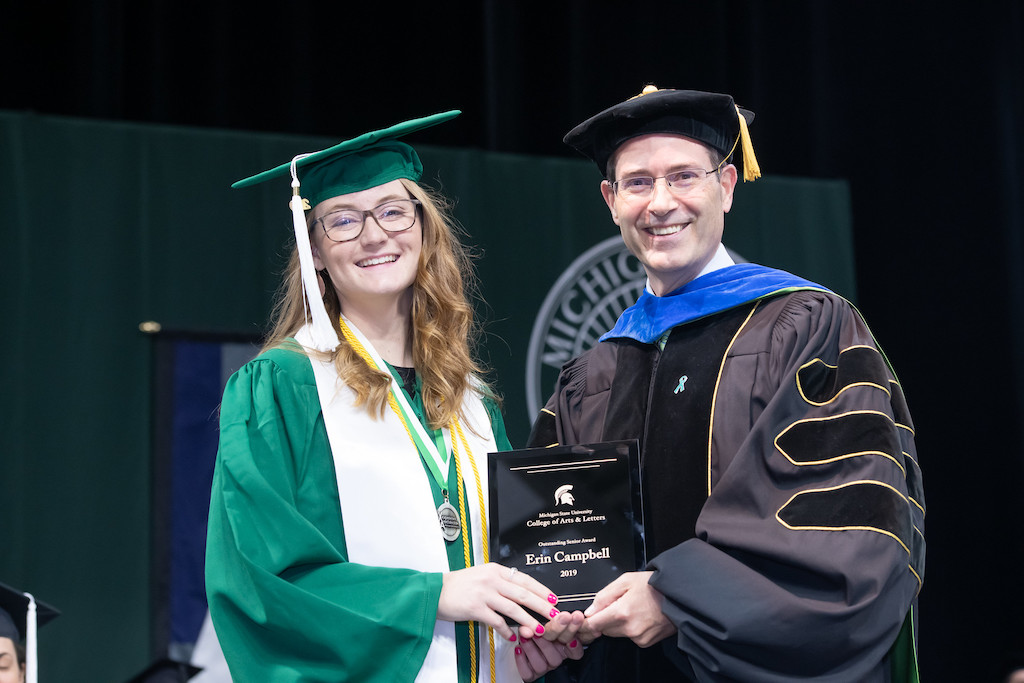 young woman who is wearing a green graduation cap and gown and man who is wearing a black graduation cap and gown who are holding a plaque in between them