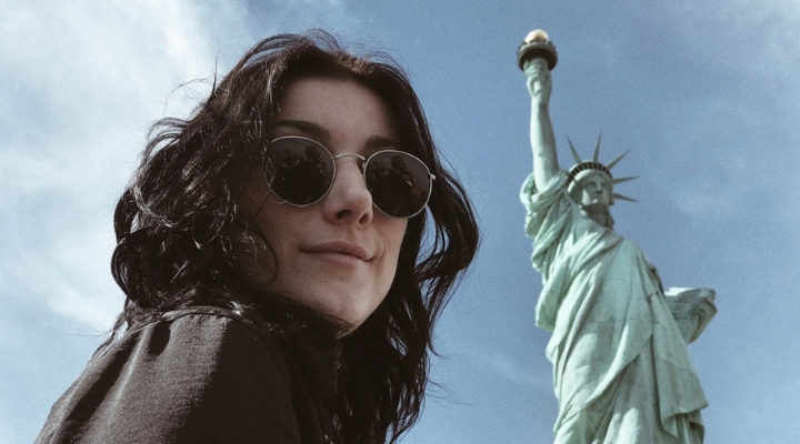 a girl with dark hair wearing sunglasses and the statue of liberty is behind her 