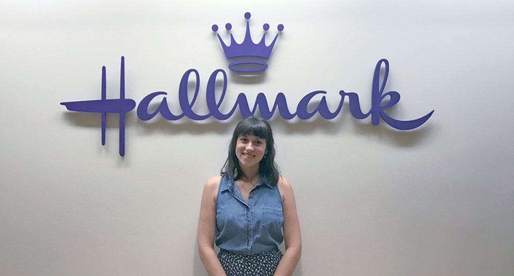 picture of a brunette woman with short hair in a denim top, she is standing in front of a wall that says 'Hallmark'