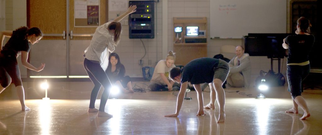 Students in various positions in front of bright lights while practicing for a performance.