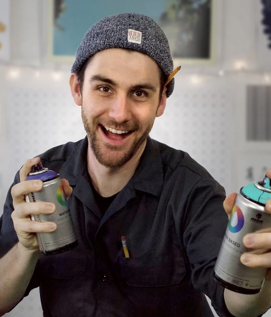 man who's wearing a black shirt and a beanie who's smiling at the camera and holding two cans of spray paint