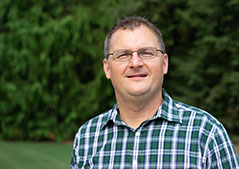 a man wearing glasses in a checkered green and blue shirt with trees behind him