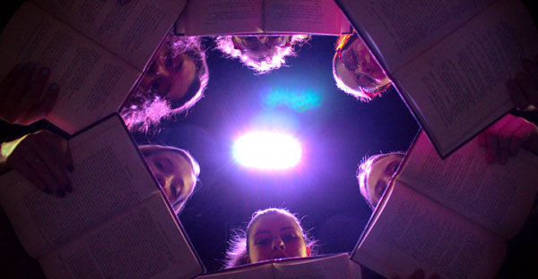 students in circle peeking over books looking down at camera w/ bright light above