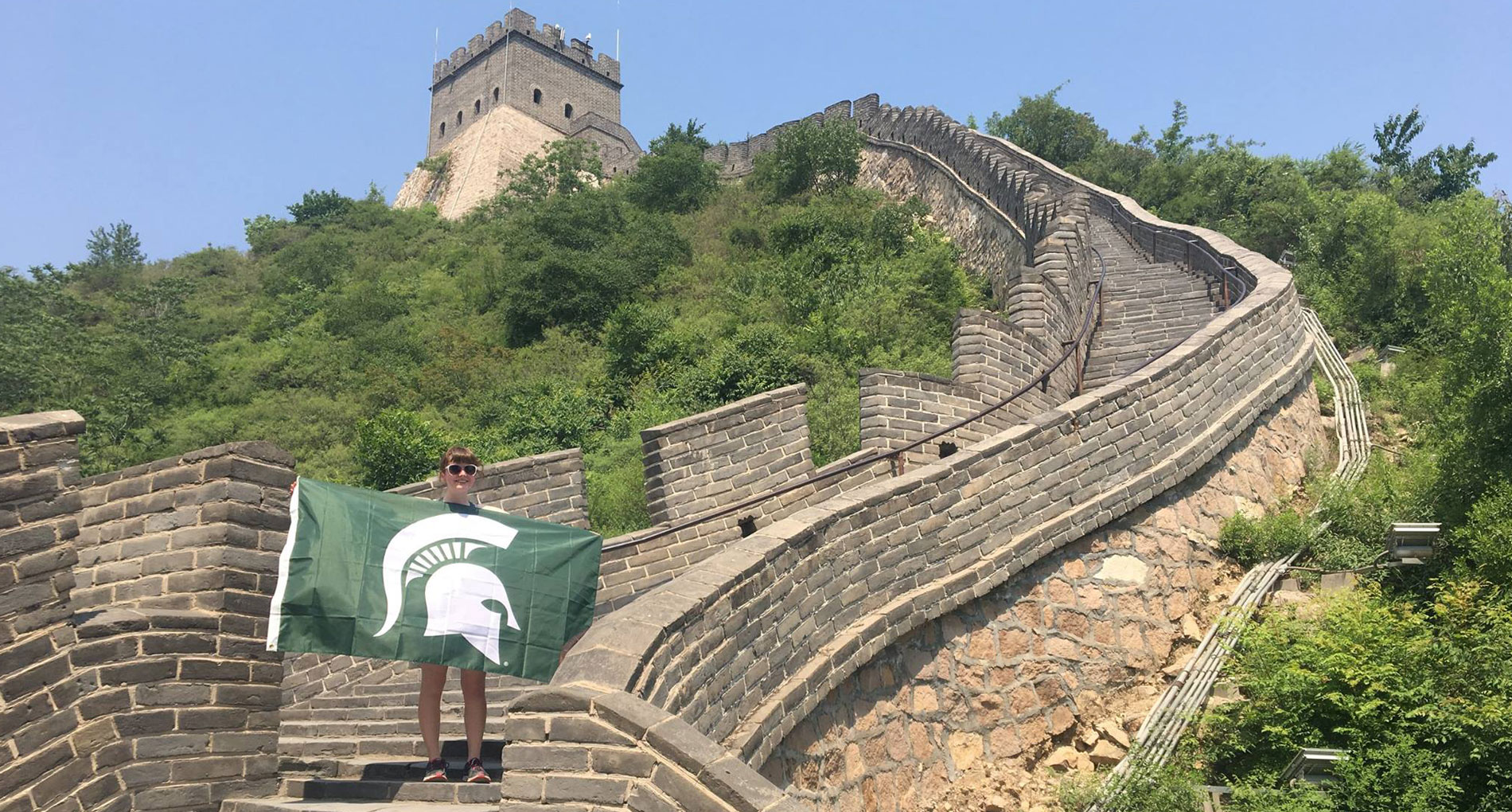 a student with tied back hair holding a spartan flag on the great wall of china
