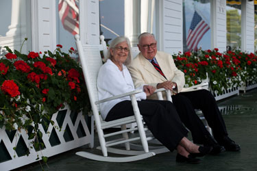 man and woman sitting on porch in white rocker chairs with red flowers surrounding them